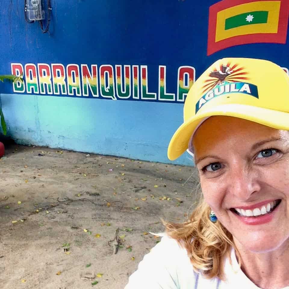A person wearing a yellow cerveza Aguila hat smiles in front of a colorful wall reading "Barranquilla," photo ©Kate Dana