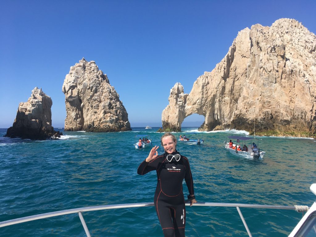 a person in a wet suit making an "OK" hand symbol stands near the famous Arco in Cabo San Lucas, Mexico during PADI Advanced Certified Diver training, photo © Kate Dana