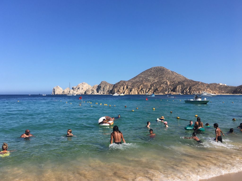 people splash in blue water with a mountain in the background of Playa Medano in Cabo San Lucas, Mexico, photo © Kate Dana