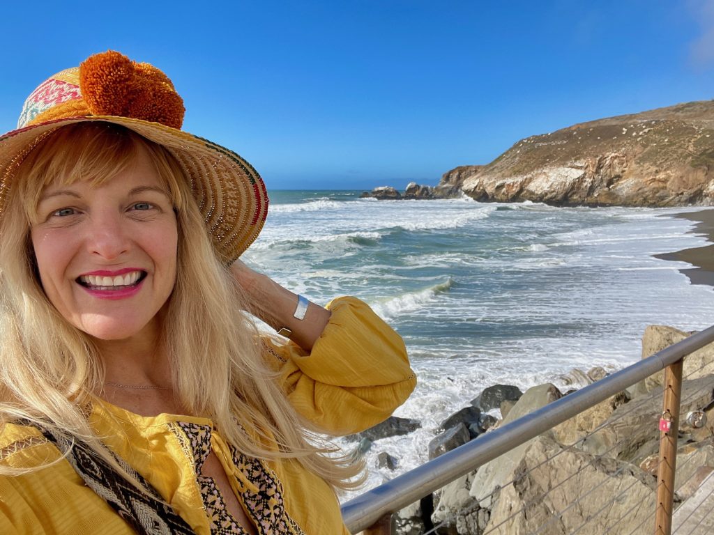 Kate Dana smiles in a yellow shirt and hat with a view of Rockaway Beach near Pacifica, CA | photo ©Kate Dana