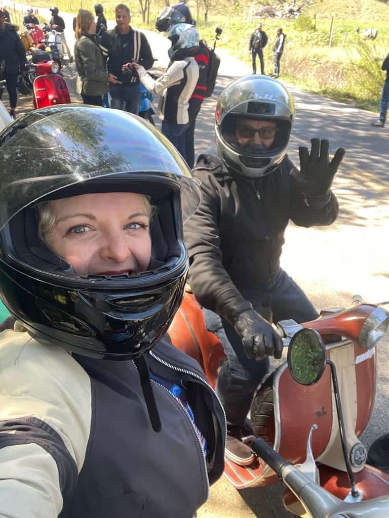 Two riders wearing helmets and riding gear on vintage Vespas, Italian motor scooters, smile for the camera at a stop with their bikes along the daytime ride route for the annual Rides of March scooter rally in San Luis Obispo, CA, photo ©Kate Dana