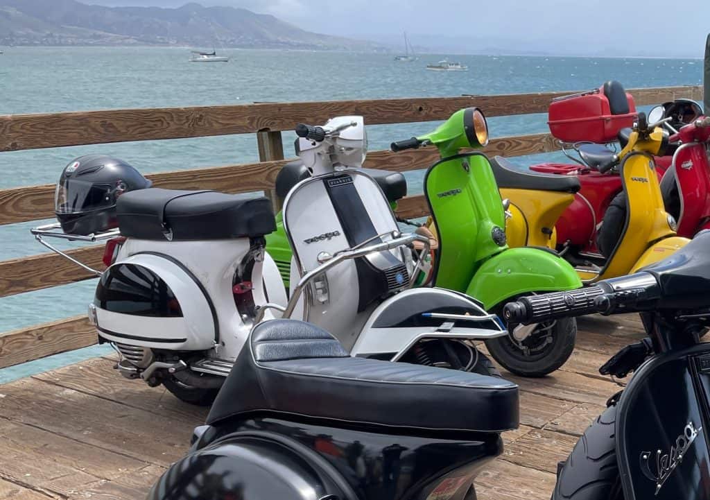 Several colorful, vintage Vespas and Lambrettas - manual-shift, Italian motor scooters - gathered for the weekend for the annual Rides of March scooter rally, sit parked side by side on the Avila Beach pier overlooking the bay and mountains near in downtown San Luis Obispo, CA, photo ©Kate Dana