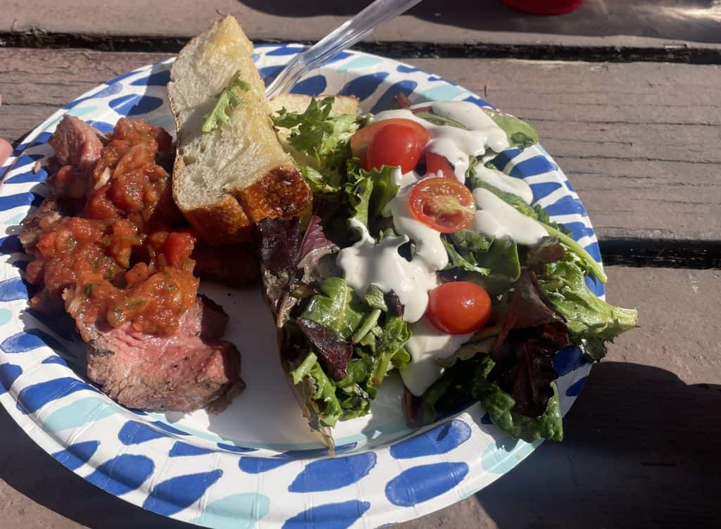 A paper plate heaped with tri-tip beef barbecue, toasted Italian bread, and a green salad with tomatoes and creamy dressing, sits on a picnic table as part of the lunch for Rides of March scooter rally in San Luis Obispo, CA, photo ©Kate Dana