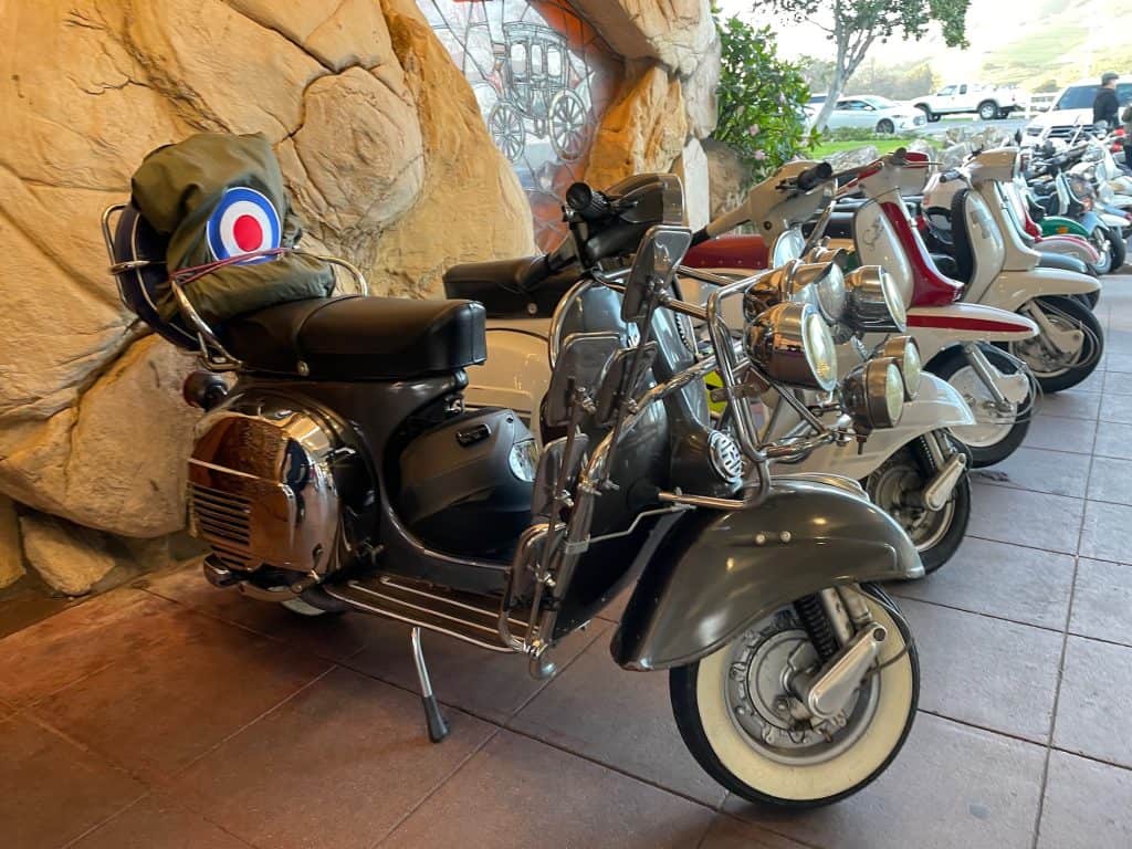A group of vintage Vespa and Lambretta Italian motor scooters with chrome trim, custom paint, and white wall tires sit parked in a line in the breezeway of the Madonna Inn, one of the gathering places for the annual Rides of March scooter rally, in San Luis Obispo, CA, photo © Kate Dana