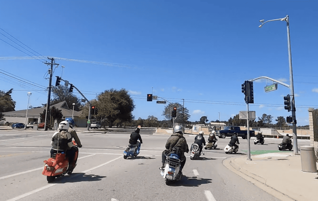 Scooterists on vintage Vespas and Lambrettas, Italian 2-stroke motor scooters, wearing riding gear and helmets, depart on a city street during a clear day for the Saturday ride during the Rides of March vintage scooter rally in San Luis Obispo, California. © Kate Dana.