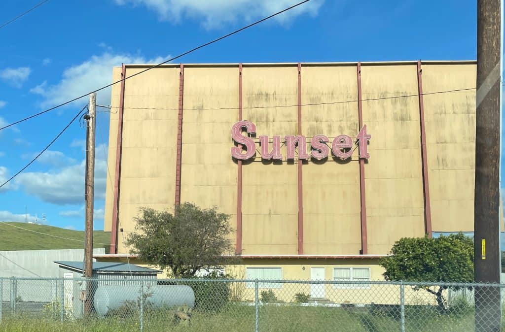 The back of the large screen at the Sunset Drive-In, near San Luis Obispo, California. The screen, against a clear blue sky, is dull yellow with the neon word "Sunset" in red, © Kate Dana
