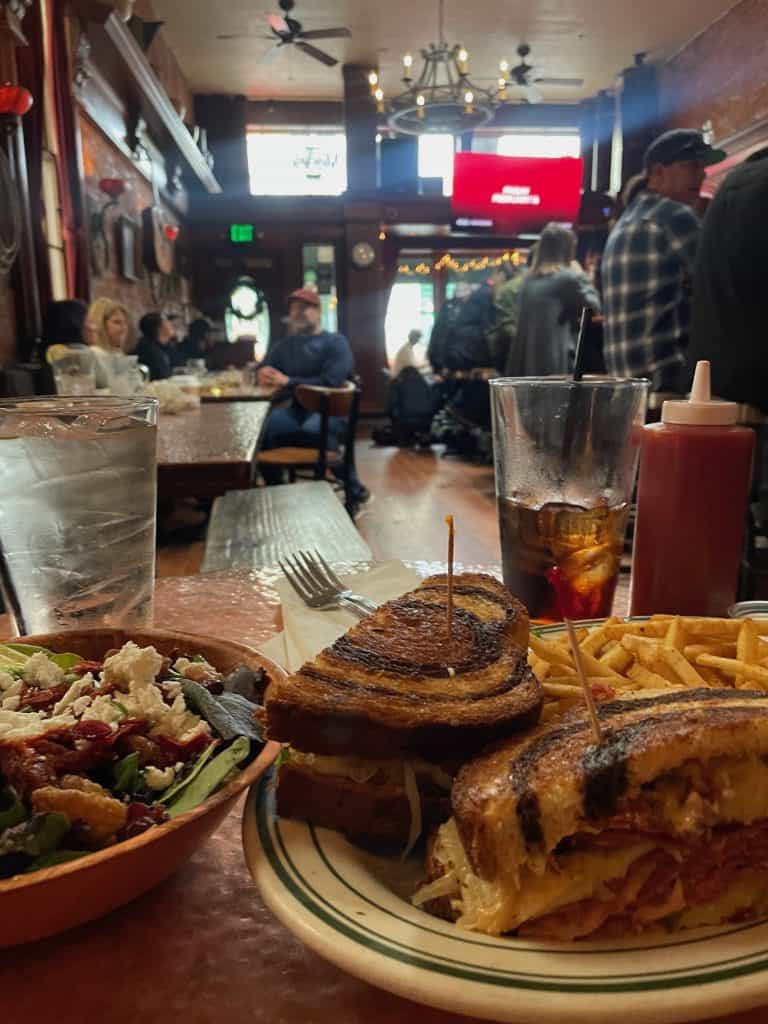 A savory mixed greens salad and a large, toasted pastrami sandwich with French fries, next to pint glasses half-filled with beverages, occupy a small table looking out to a crowd of patrons in the Black Sheep Bar and Grill, in San Luis Obispo, CA