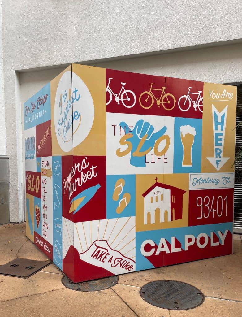 A large cube painted in gold, pale blue, burgundy, and white announcing the unique features of the town like biking, Cal Poly, and the "SLO life," fills a space on a sidewalk in downtown San Luis Obispo, CA, location of the annual Rides of March scooter rally, photo ©Kate Dana