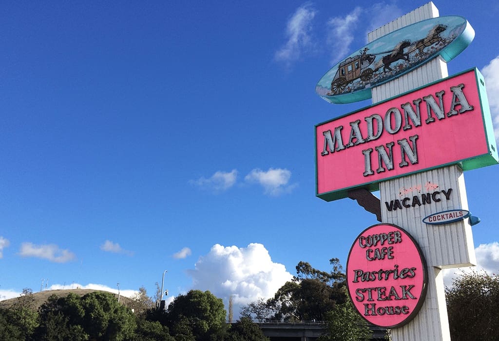 A large, pink-and-white neon sign for the Madonna Inn, one of the gathering places for the annual Rides of March scooter rally, beams against a sunny, blue sky featuring a horse and carriage graphic and announcing the Copper Cafe Pastries and Steak House in San Luis Obispo, CA, photo © Kate Dana