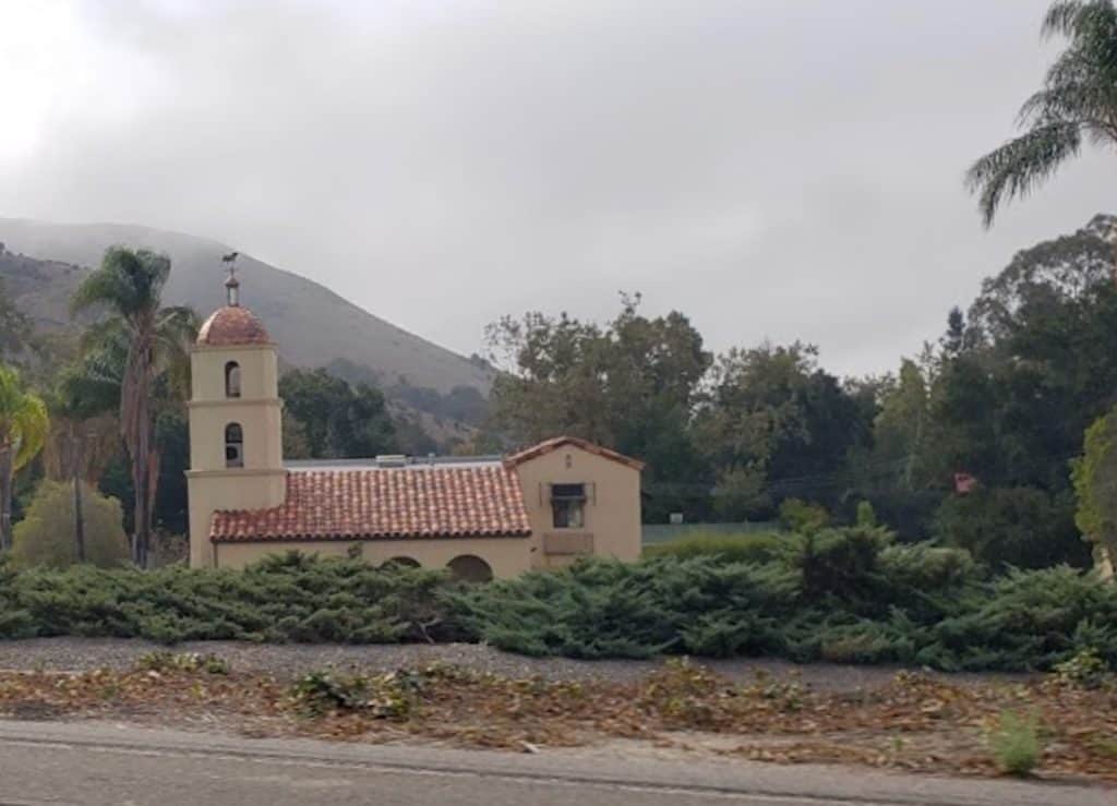 A small, Spanish style building with a terra cotta tile roof and bell tower occupies a small lot on the side of a busy highway, nestled amid trees, showing what remains of the Mo-tel Inn, the world's first motor hotel near San Luis Obispo, © Kate Dana