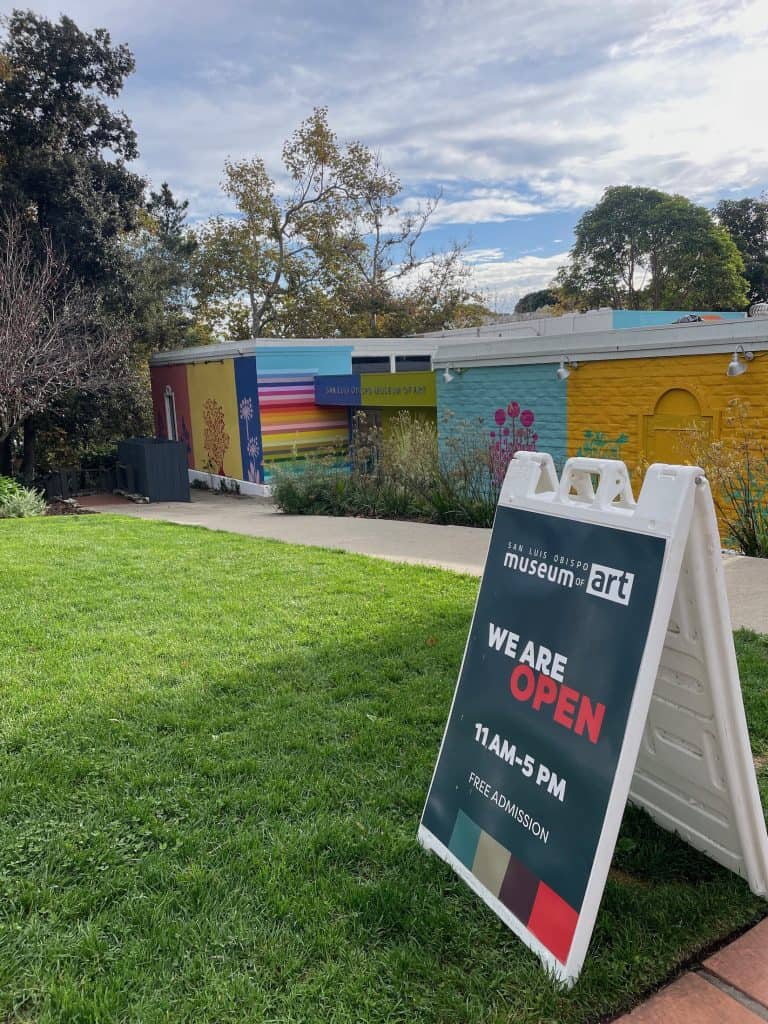 A printed sandwich-board type sign sits on a grassy lawn in front of a colorful building on a clear, sunny day, announcing that the San Luis Obispo, California Museum of Art is open 11 AM to 5 PM. © Kate Dana