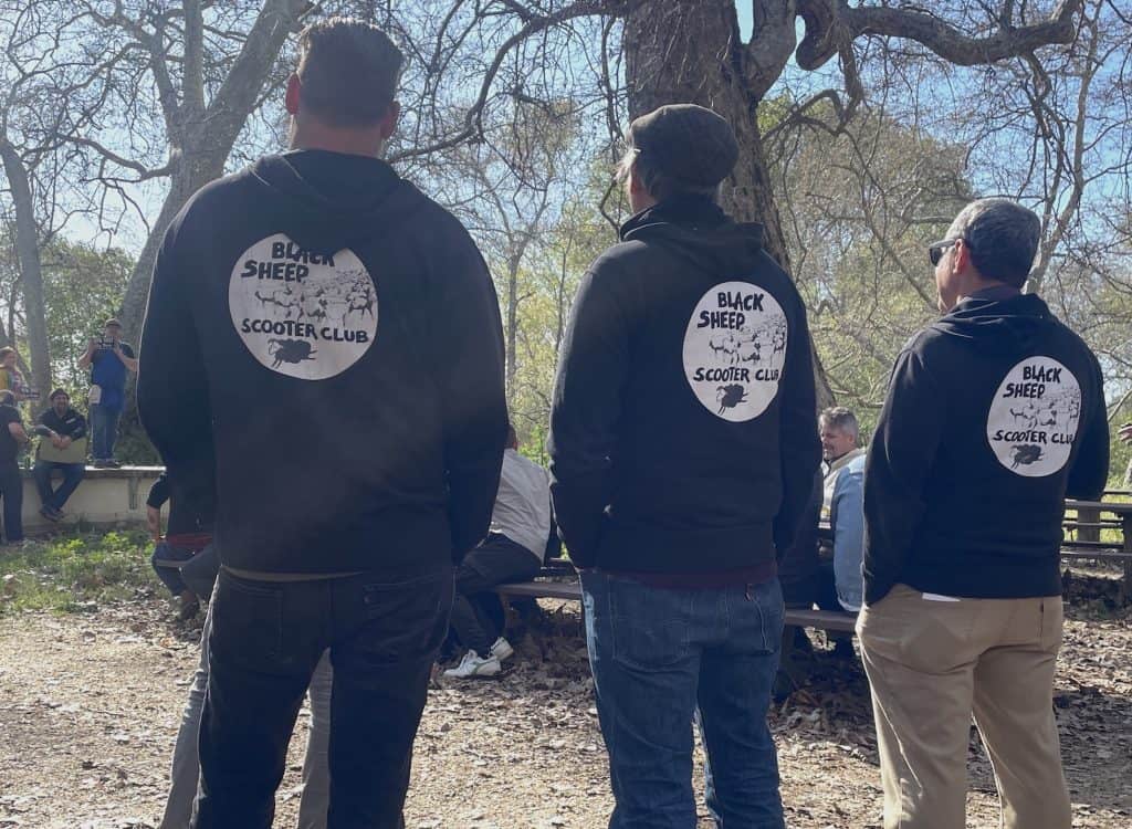 Three members of the Black Sheep Scooter Club from San Francisco, CA, wearing their club patches on the back of their black jackets, stand in a park facing an announcer during the raffle for the annual Rides of March scooter rally held in San Luis Obispo, CA, photo ©Kate Dana