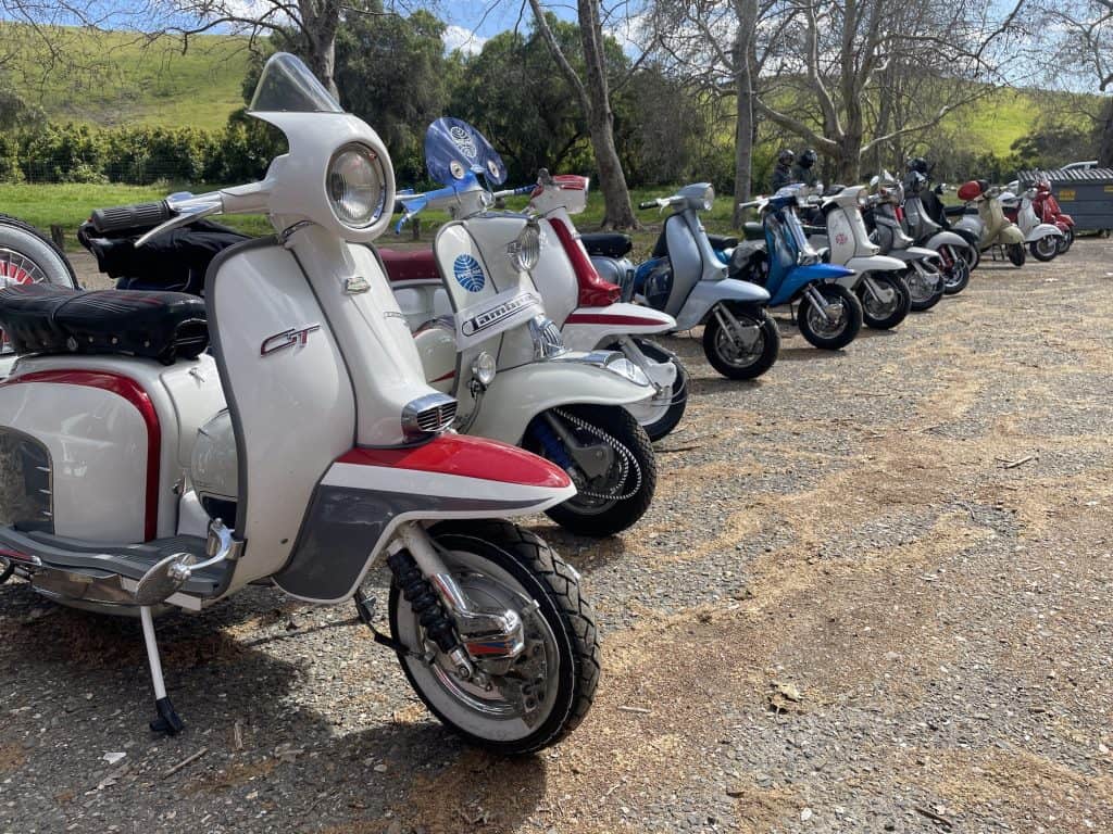 Colorful, vintage Lambrettas and Vespas, Italian-made motor scooters visiting San Luis Obispo, California, for the annual Rides of March rally, sit parked side-by-side in the lot of a day-use public park, photo ©Kate Dana