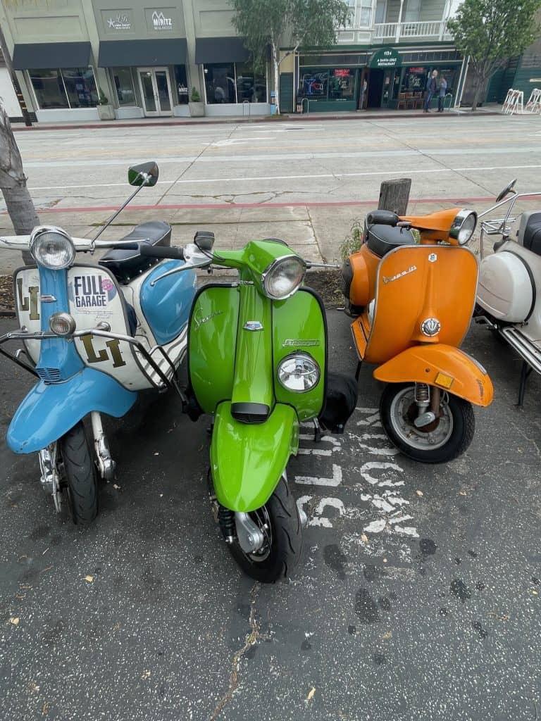 Two colorful, vintage Lambrettas and a bright orange vintage Vespa park side-by-side n a parking lot, waiting for the ride to start for the Morning Wood Classic Scooter Weekend in 2022, photo ©Kate Dana