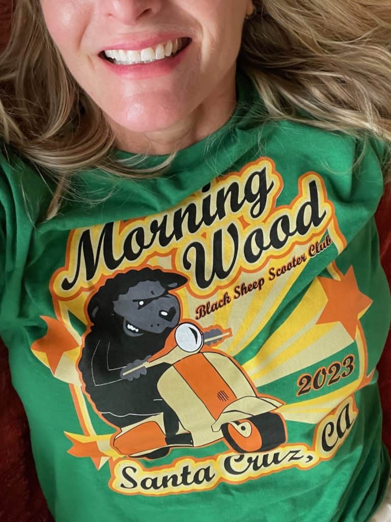 A close-up of a person wearing a colorful, green t-shirt with 2023 artwork in orange, yellow, black and grey featuring a graphic by Phil Carey of a sheep riding a vintage scooter commemorates of Morning Wood Classic Scooter Weekend in Santa Cruz, CA photo ©Kate Dana