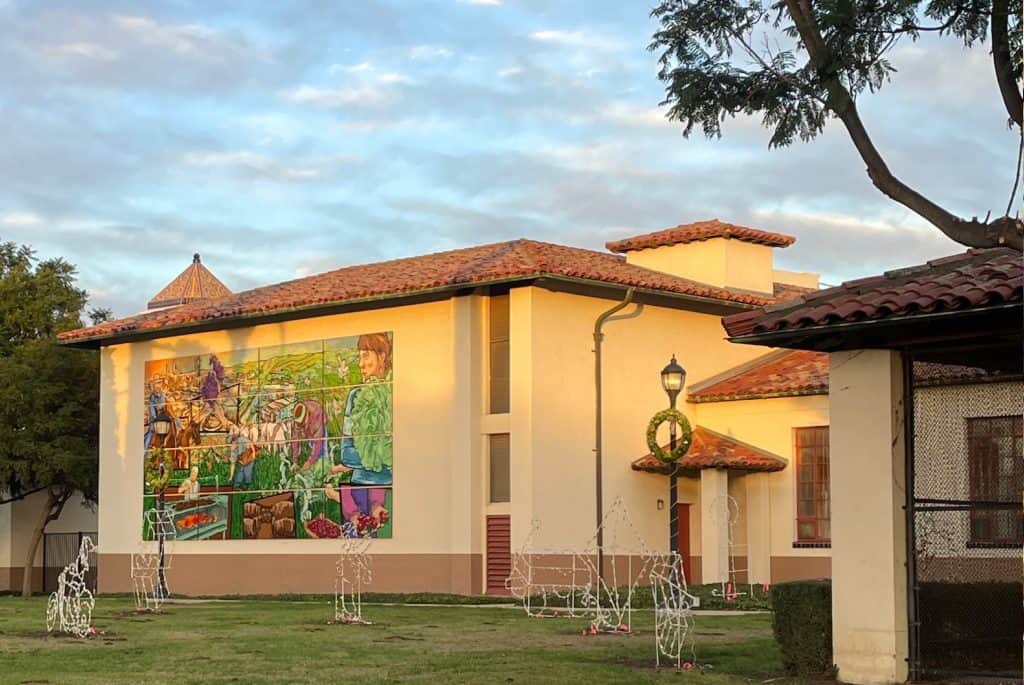 The Santa Maria community center, a small brick building with a colorful tile mural on one wall, sits in the center of a grassy park with a blue sky and clouds overhead, photo © Kate Dana.