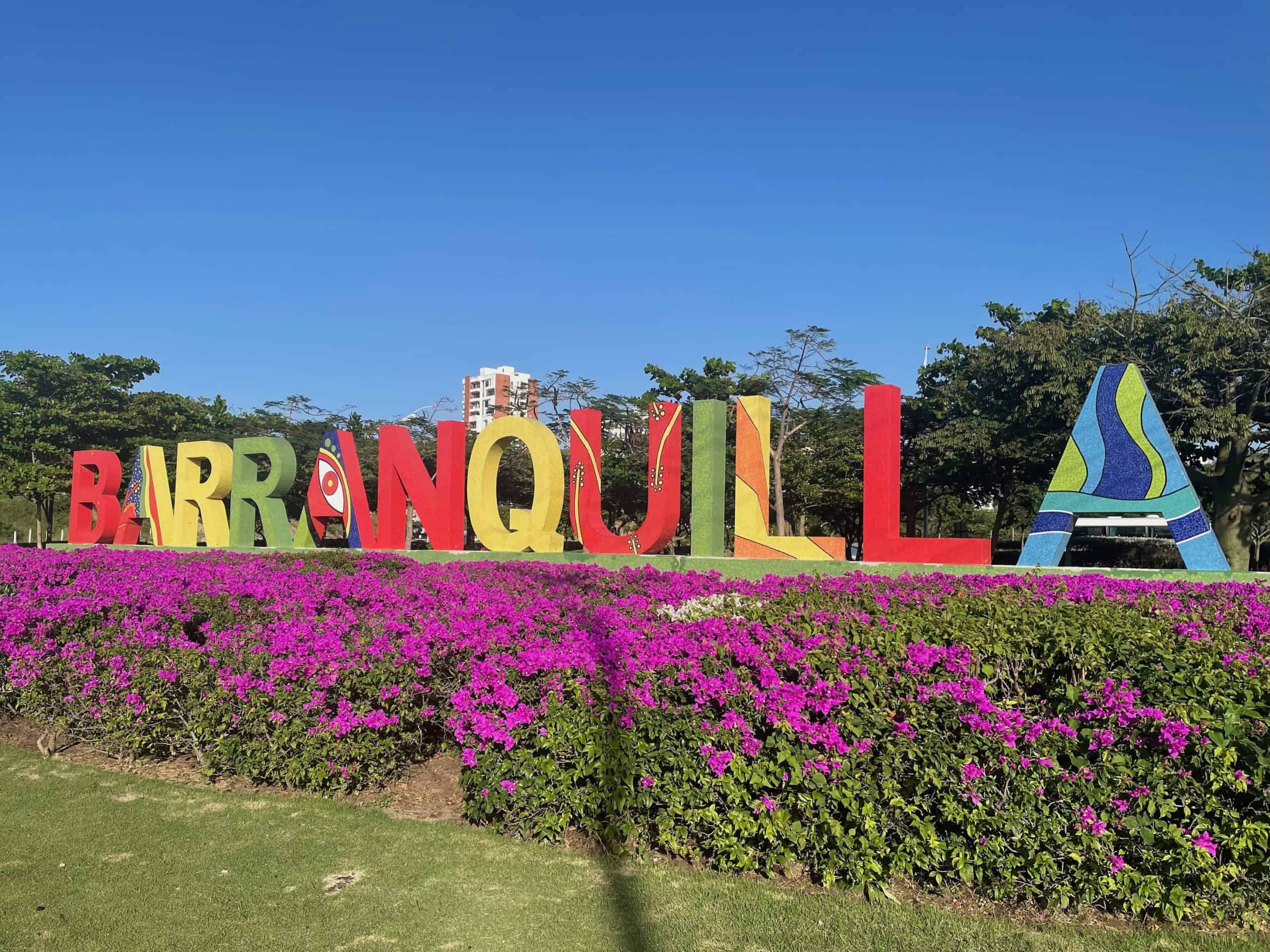 Large, colorful letters spell out the city of Barranquilla in Colombia against a grassy field with pink flowers in the foreground in this photo ©Kate Dana