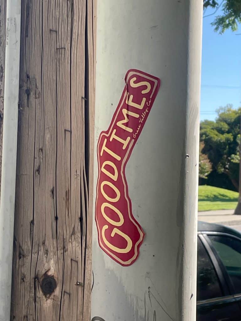 A red and cream-colored sticker in the shape of the state of California stuck to an electrical pole reads "Good Times" in this close-up photo for 45 California Sayings to Get Stoked About Right Now, photo © Kate Dana