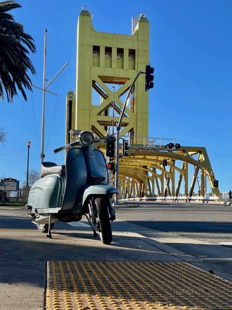A light blue, 1969 Lambretta motor scooter sits on a sidewalk with the yellow Tower Bridge in the background behind it, on a sunny day in this photo for 45 California Sayings to Get Stoked About Right Now, photo © Kate Dana