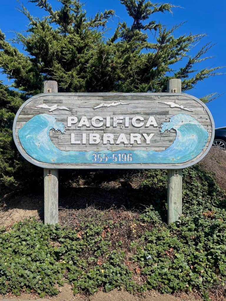 A salt-and-sea weathered sign announcing the Pacifica Library with waves carved into it posts up in front of a green shrub on a sunny day with clear skies in this photo for 45 California Sayings to Get Stoked About Right Now, photo © Kate Dana