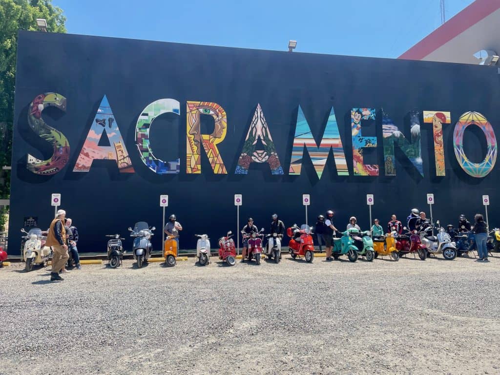 Several vintage and modern Vespa and Lambretta motor scooters park next to each other, lined up in front of a black building with a colorful mural reading, "Sacramento" in this photo for 45 California Sayings to Get Stoked About Right Now, photo © Kate Dana