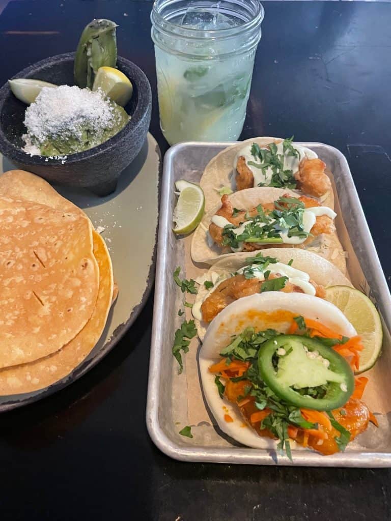 Delicious homemade tacos and a plate of tostadas next to a molcajete with green salsa on a black shiny table next to an icy lemonade, photo © Kate Dana
