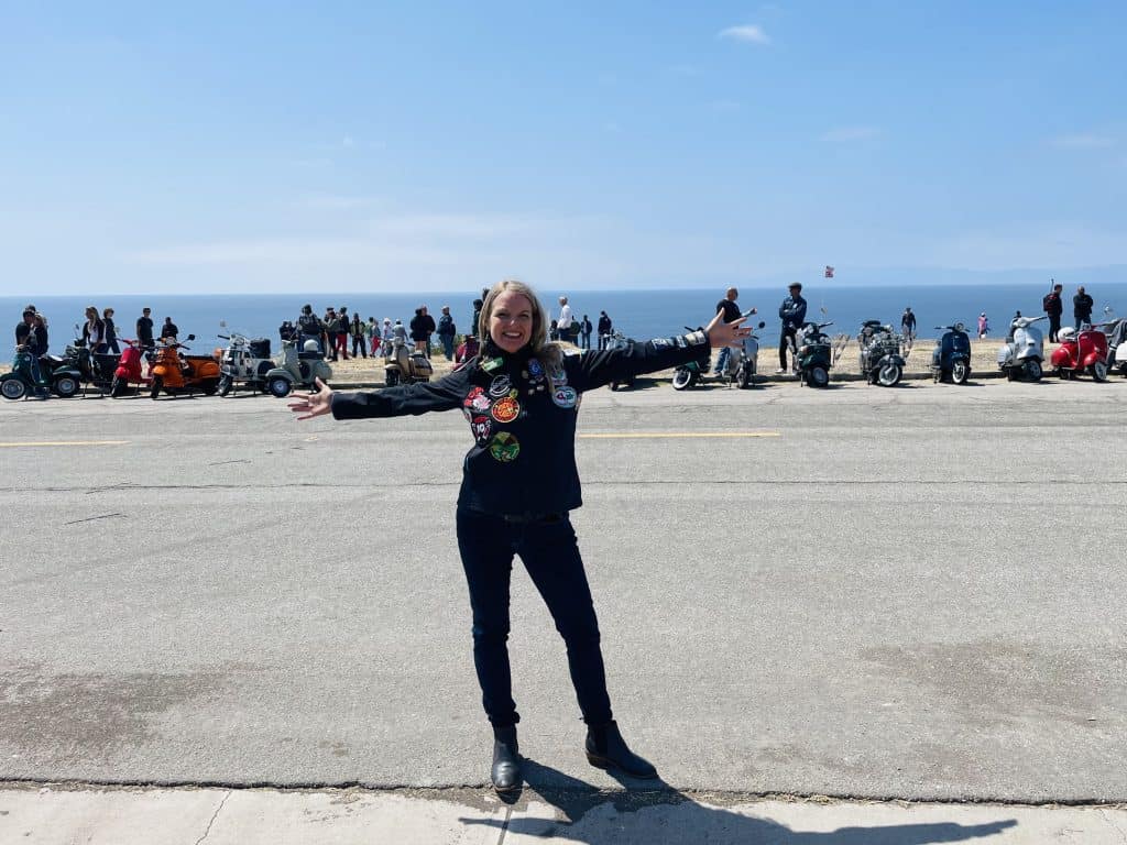 Kate Dana stands with her arms stretched wide, wearing a patch-covered jacket, across the street from dozens of vintage Vespas and Lambrettas, lined up overlooking the ocean near Long Beach, CA, home of Long Beach Classic Scooter Club's annual Classic Not Plastic Scooter Rally on Memorial Day weekend, photo ©Kate Dana