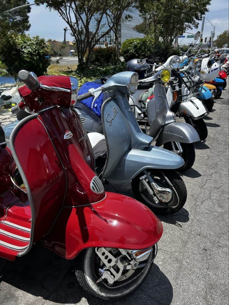 A red vintage Lambretta, a blue Lambretta, and several other scooters parked side by side on a street curb, photo ©Kate Dana