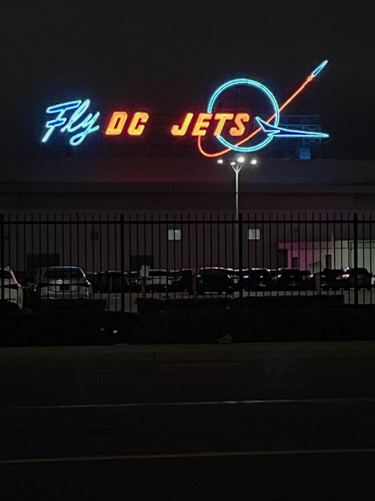 A red and blue neon sign announcing Fly DC Jets with a circular logo and airplane silhouette, glows in the night sky near Long Beach airport in California, home of Long Beach Classic Scooter Club's annual Classic Not Plastic Scooter Rally on Memorial Day weekend, photo ©Kate Dana