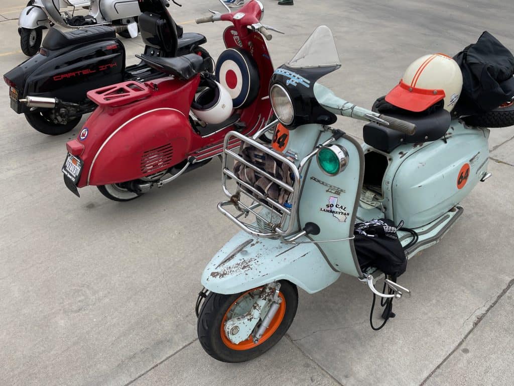 A close up image of a red, vintage Vespa sits parked between two vintage Lambrettas, one featuring Cartel Ink, in a parking lot in Long Beach, CA, home of Long Beach Classic Scooter Club's annual Classic Not Plastic Scooter Rally on Memorial Day weekend, photo ©Kate Dana