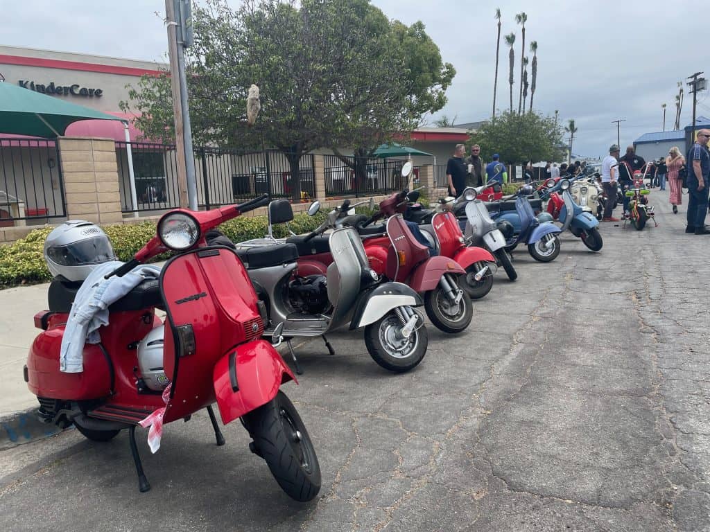 Dozens of vintage Vespas and Lambrettas, sit parked, lined up near a sidewalk in Long Beach, CA, home of Long Beach Classic Scooter Club's annual Classic Not Plastic Scooter Rally on Memorial Day weekend, photo ©Kate Dana