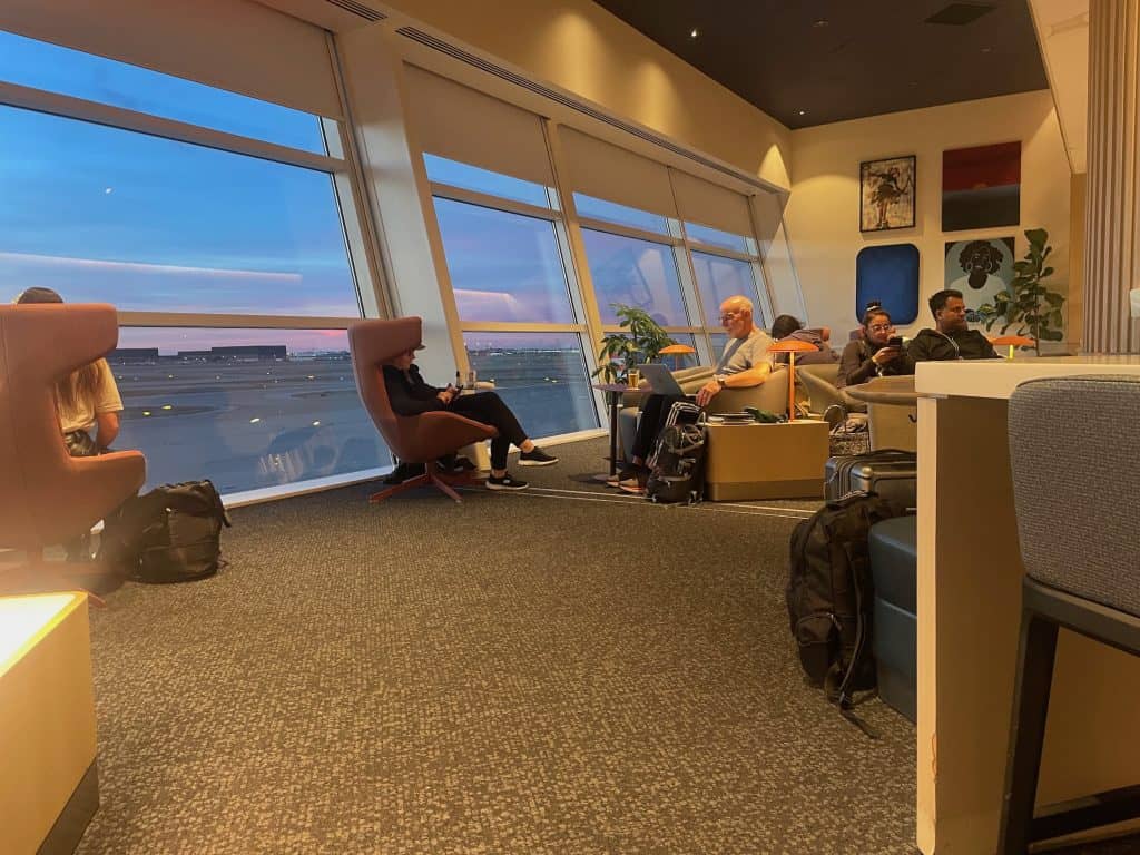 An inside view of soft, large chairs in front of a huge picture window as the sunsets over the tarmac in Texas. The Capital One Lounge in Dallas Fort Worth (DFW) airport provides comfortable seating, free WIFI, complimentary food and drinks, showers, nap rooms, and more to approved members and is available as part of the Priority Pass program, photo ©Kate Dana