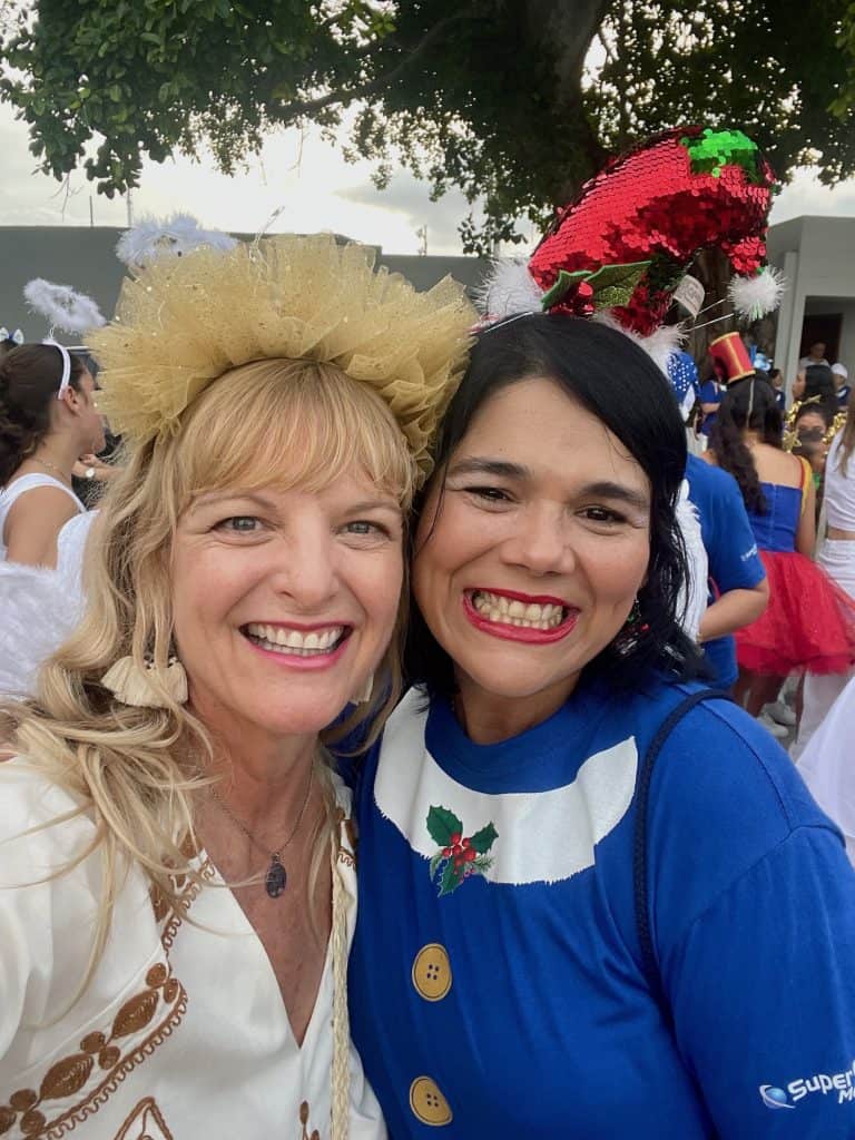 A blonde person smiles next to a person with dark hair in this outdoor image captured during a parade in Cartagena, Colombia, "cara a cara," face to face or cheek to cheek, they smile warmly at the camera in this photo ©Kate Dana