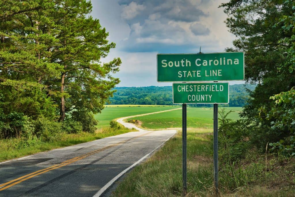 A green roadside sign announcing the South Carolina state line in Chesterfield county rises up from a grassy field before long, winding country roads in this color, close-up photo for 78 Southern Phrases to Blow Your Hair Back, Cocameca.com photo © Clint Patterson for Unsplash