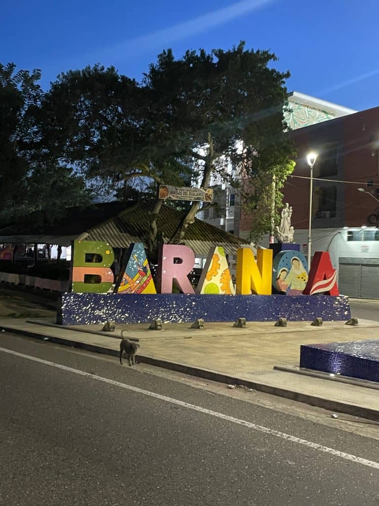 A large, colorful sign announcing the town of Baranoa in yellow, orange, blue, and red sits in a median between a busy street against a clear blue sky during a day en route to enchanting Usiacurí, Colombia, photo ©Kate Dana