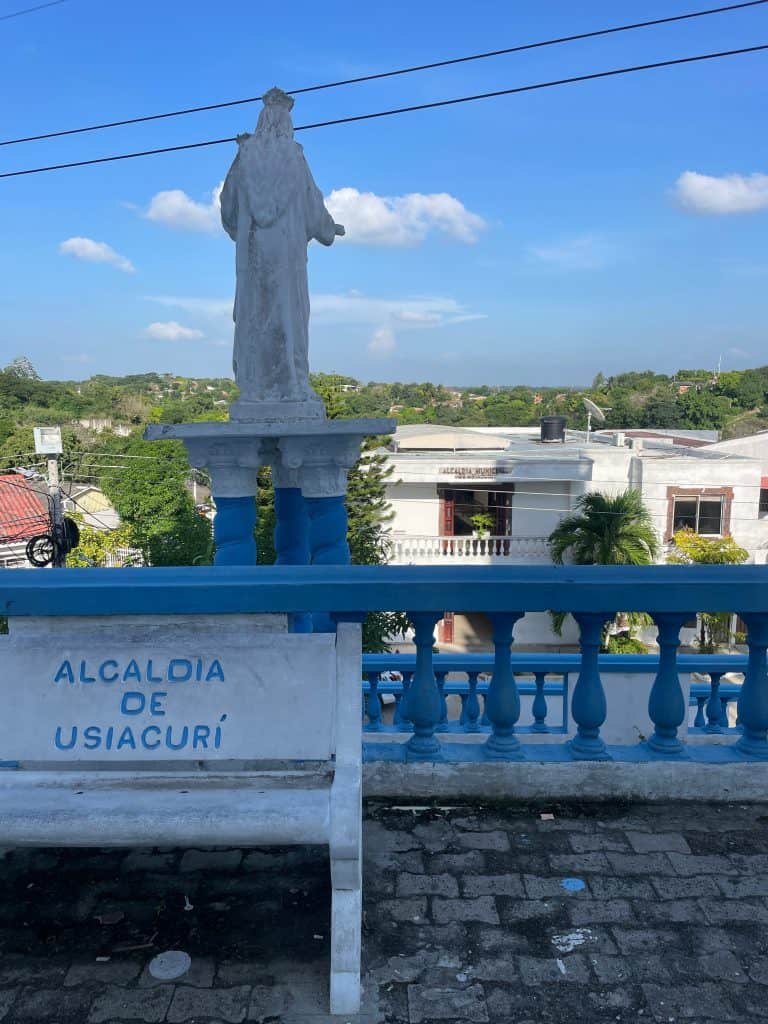 A white angelic statue overlooks the street below as a white bench with blue lettering announces Alcaldía de Usiacurí in front of Iglesia de Santo Domingo de Guzman during this day in enchanting Usiacuri, Colombia, photo ©Kate Dana