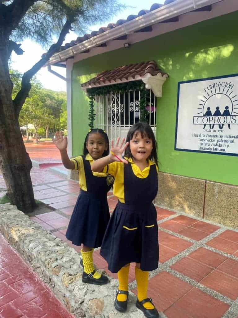 Two, friendly young children in blue and yellow school uniforms stand on a roadside and wave enthusiastically next to Fundación Coprous during a day in enchanting Usiacuri, Colombia, photo ©Kate Dana