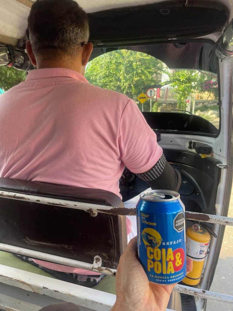 A view from the rear seat of a motocarro shows a hand holding Cola y Pola, a refajo beverage in a blue and yellow can, while a driver steers the vehicle carefully during a day in enchanting Usiacuri, Colombia, photo ©Kate Dana