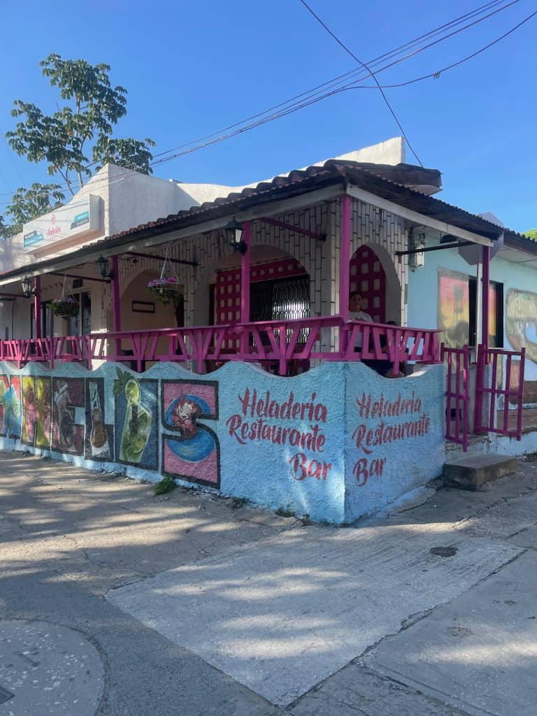 A pink balcony with terra cotta roof announces Heladeria Restaurante Bar and frutería Anhelos on a roadside below a bright blue sky during a day in enchanting Usiacuri, Colombia, photo ©Kate Dana
