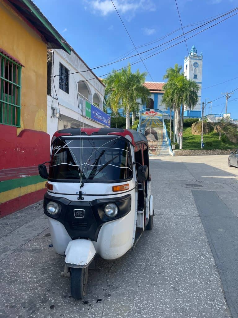A white motocarro with black trim and darkened windows waits in the street with Iglesia de Santo Domingo de Guzman rising in the background against a clear blue sky during a day in enchanting Usiacurí, Colombia, photo ©Kate Dana