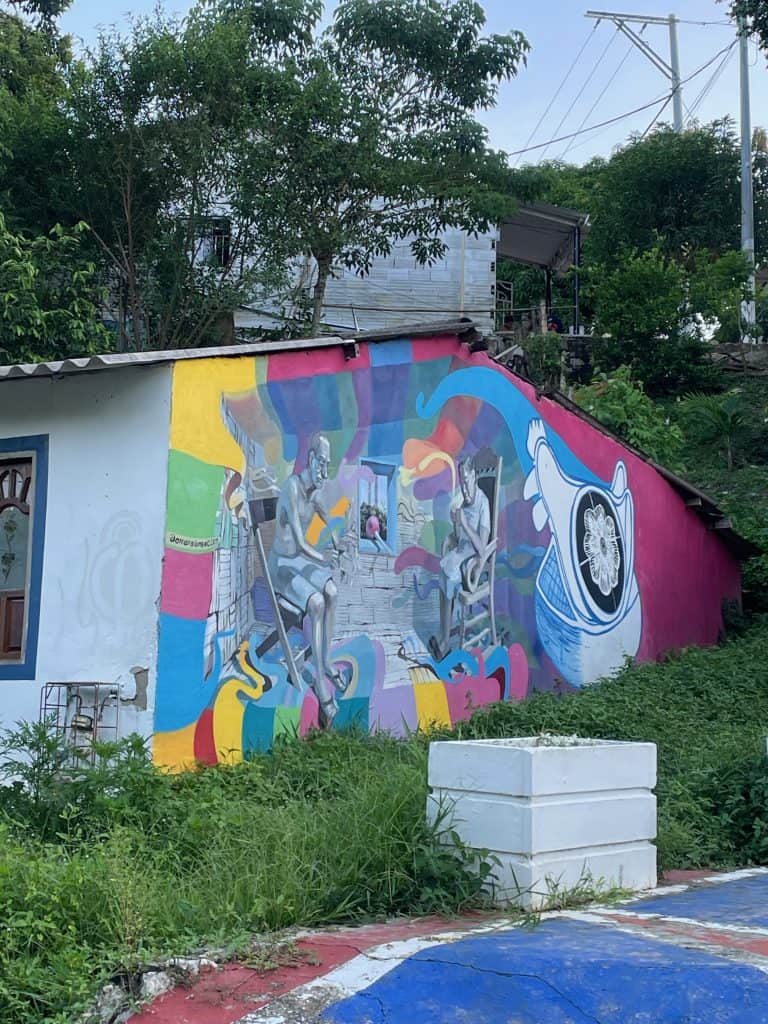 A multicolored mural with pink, yellow, and blue swirls painted on a wall of a building next to a grassy space during a day in enchanting Usiacuri, Colombia, photo ©Kate Dana