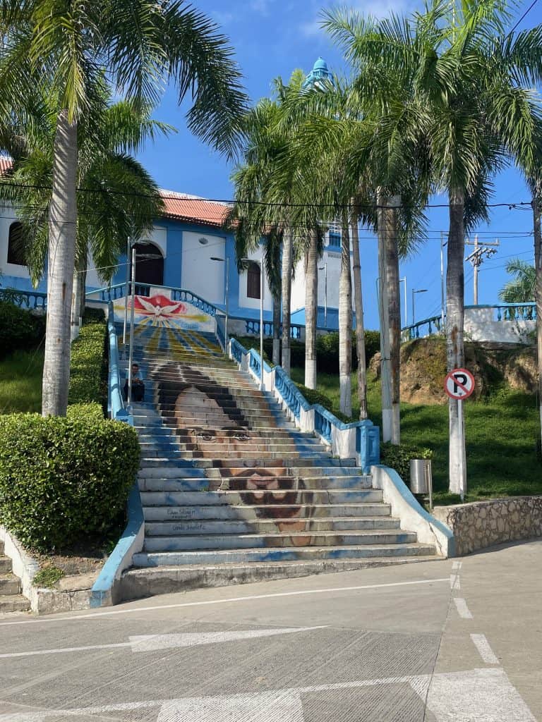 The steps leading up to Iglesia de Santo Domingo de Guzman feature the face of Jesus Christ painted cleverly onto the stairs rising from the street to the church with mature palm trees against a clear blue sky during a day in enchanting Usiacurí, Colombia, photo ©Kate Dana