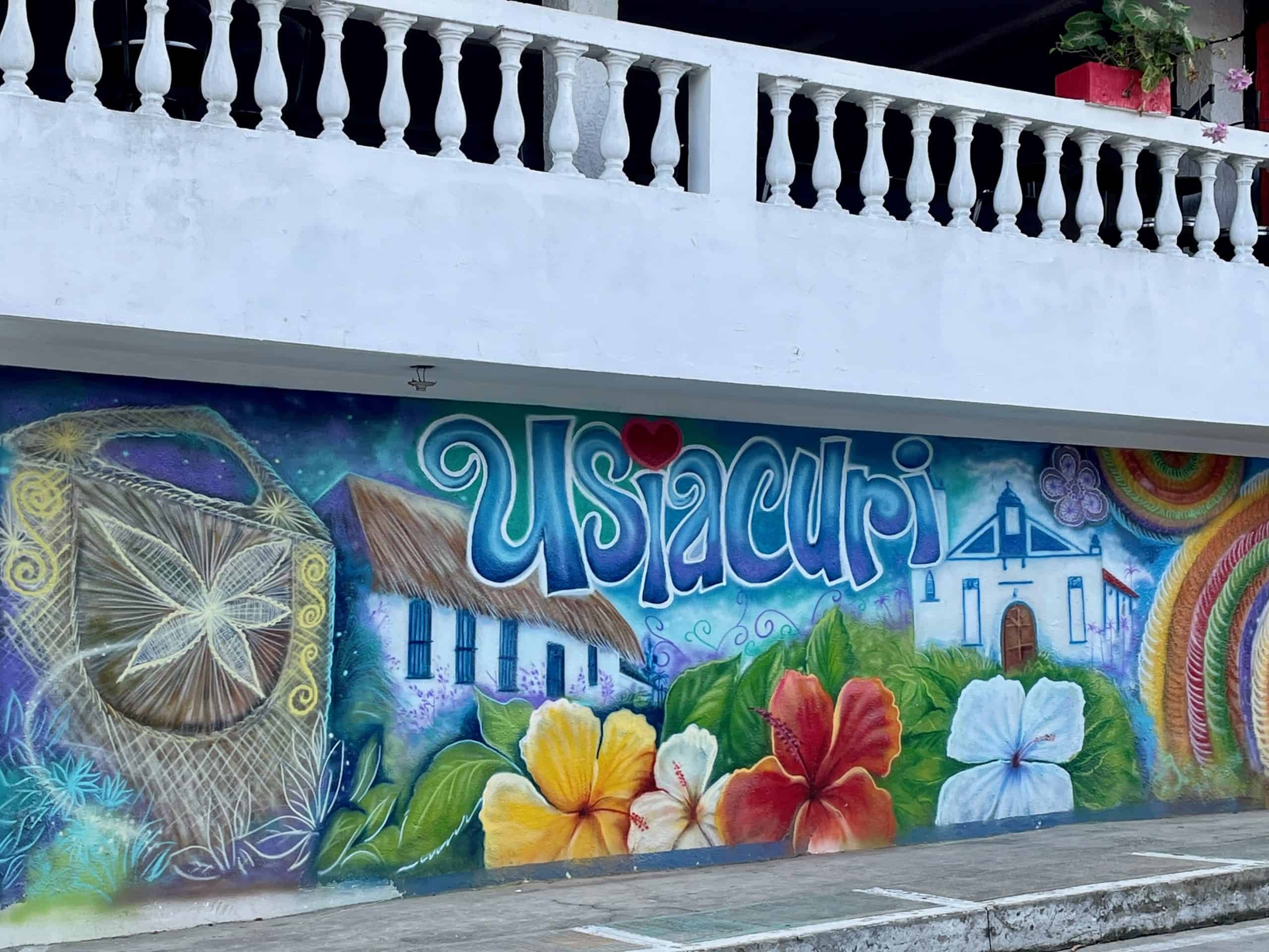 A multicolored mural on the side of a building announcing "Usiacurí" in script letters with large red and yellow flowers cover a wall beside a street in this photo taken in Usiacuri, Colombia, photo ©Kate Dana