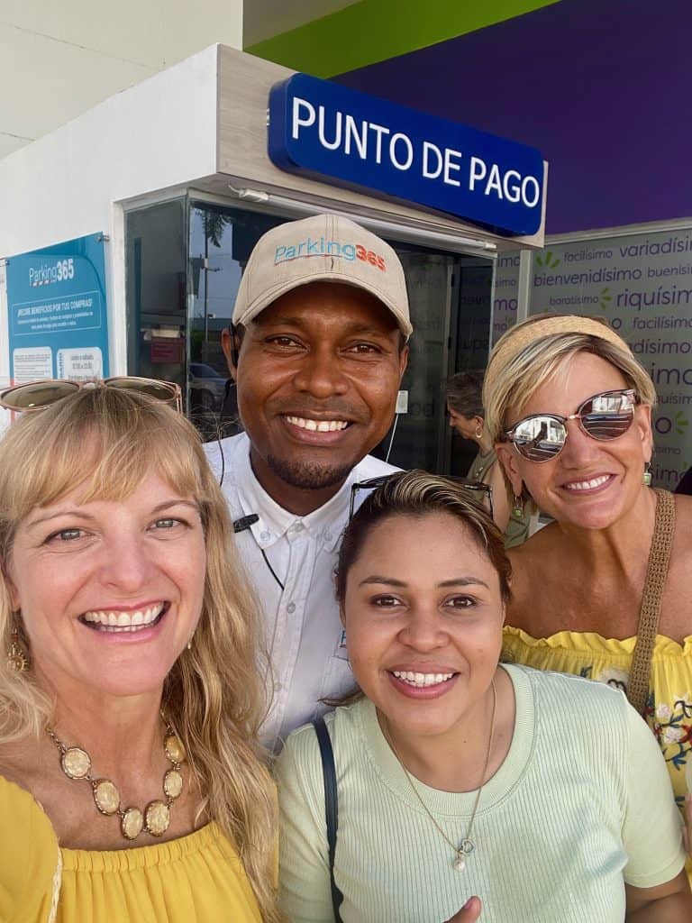 Four people with contrasting skin tones, bright smiles, and casual clothing, smile collectively at the camera with a small booth in the background that reads "punto de pago" in this close-up photo taken in Cartagena, Colombia by ©Kate Dana