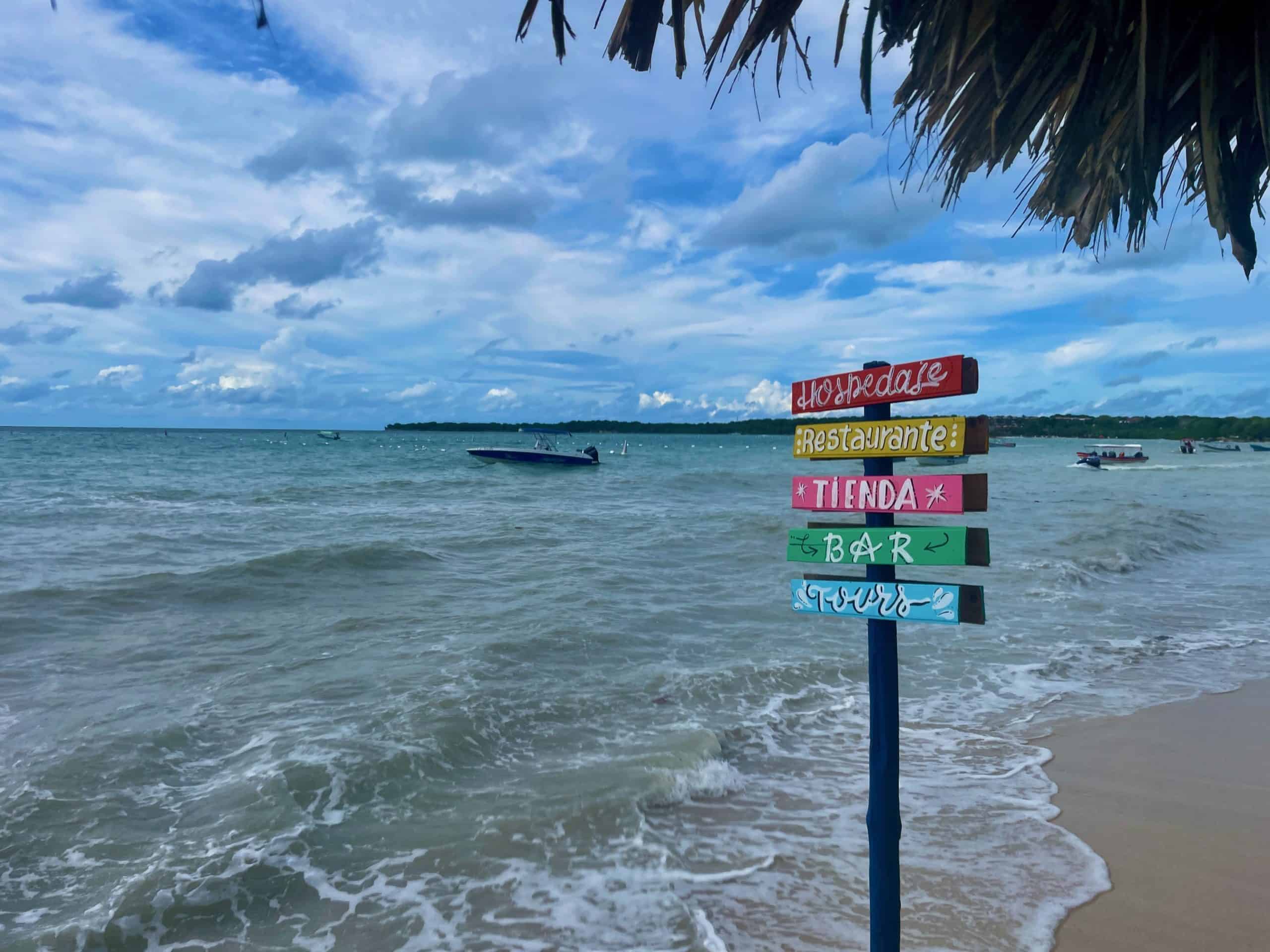 Discover authentic experiences. Embrace new adventures. On a remote, sunny beach, a colorful sign in Spanish reads: hospedaje, restaurante, tienda, bar, tours. Photo ©Kate Dana