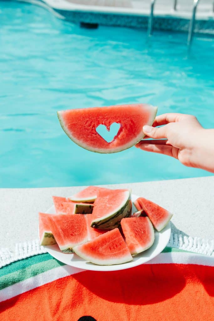 A heart cut out of a piece of watermelon being held 
by a hand in front of a swimming pool in this color photo for 78 Southern Phrases to Blow Your Hair Back, Cocameca.com photo ©Melton Kenta Kikuchi for Unsplash