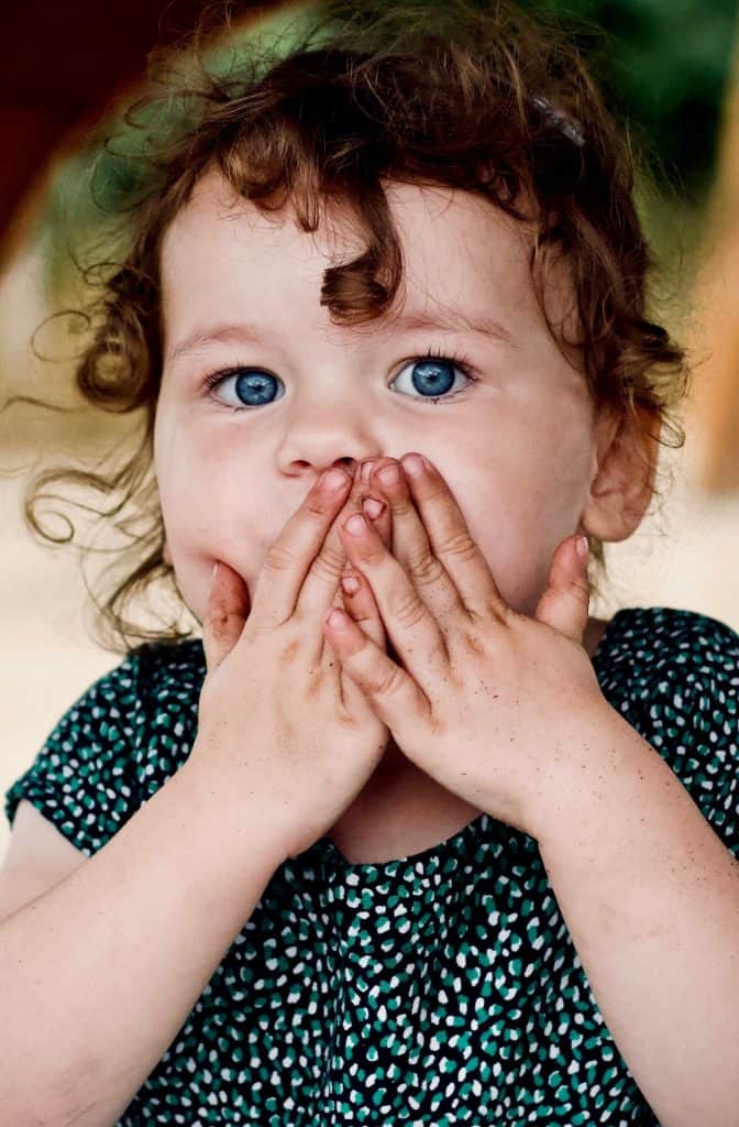 "Hush, now": A child with big blue eyes and curly brown hair holds her hands over her mouth in surprise in this color, close-up photo for 78 Southern Phrases to Blow Your Hair Back, Cocameca.com photo © Jelleke-Vanooteghem for Unsplash