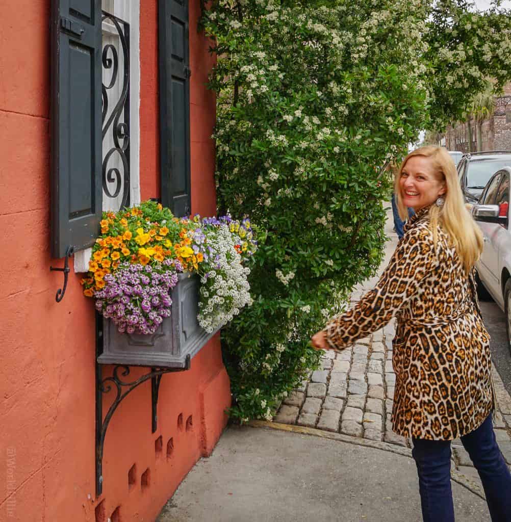 "Hey, y'all!" Kate Dana looks over her shoulder wearing a leopard print coat and dark blue jeans next to a terra-cotta colored house with purple and yellow flowers in windowboxes during a walk in Charleston, S.C, in this color, close-up photo for 78 Southern Phrases to Blow Your Hair Back, Cocameca.com photo © Kate Dana