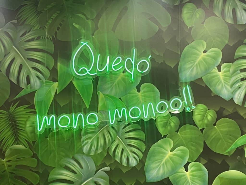 A green neon sign reads Quedó, mono monooo!! against green leaves in this photo from Colombia by ©Kate Dana