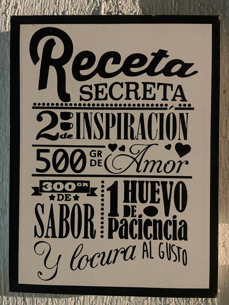 A sign tells the Receta Secreta of 2 cups of inspriación, 500 grams of amor, 300 grams of sabor, 1 huevo (egg) of patience, and whip with gusto in this black and white wall sign photographed in Colombia by ©Kate Dana
